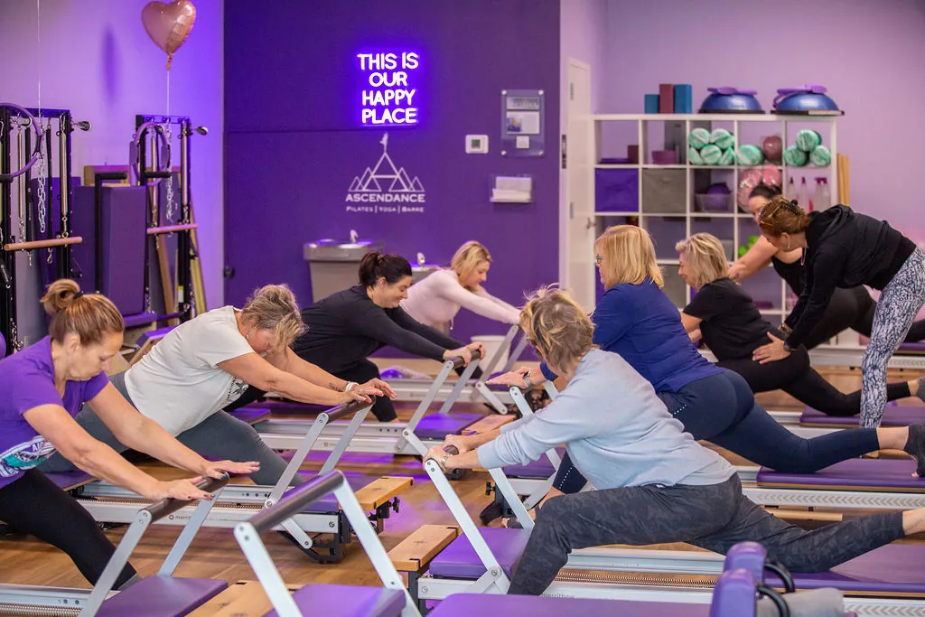 A Reformer Pilates class at Ascendance stretching together under the studio's This Is Our Happy Place neon sign